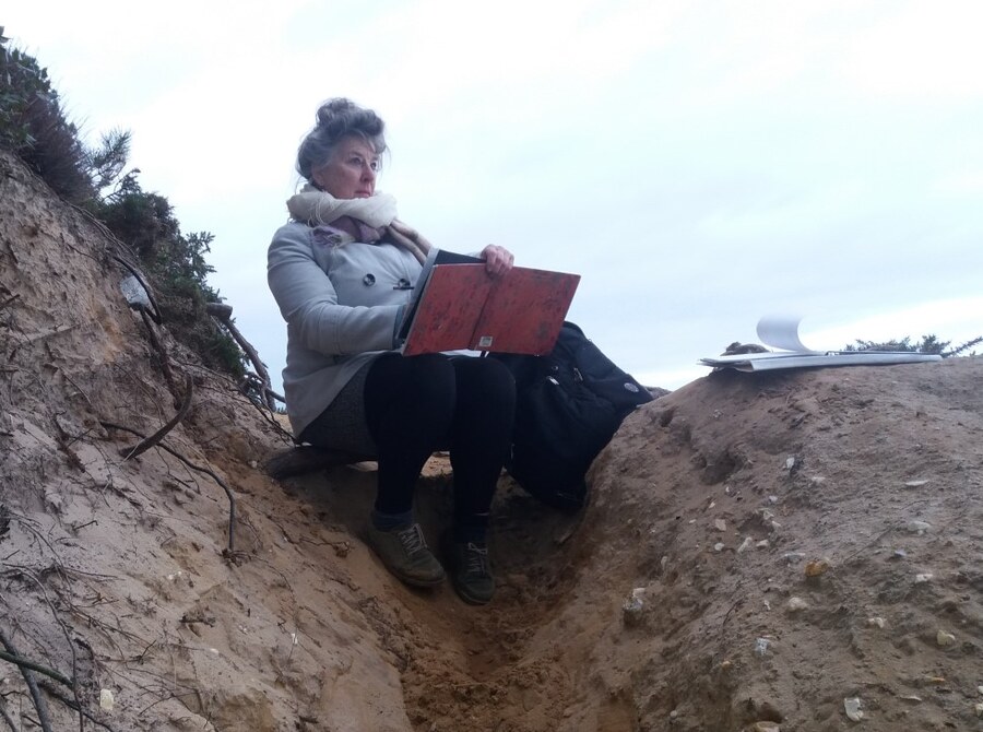 Lizzy Short drawing outdoors, seated in a sandy cliff gully with a board on her knees